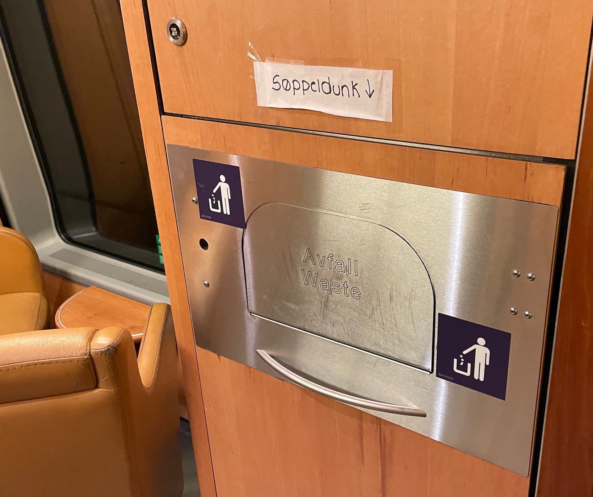 En søppeldunk på et tog. Over dunken er det hengt opp en papirlapp hvor det står "Søppeldunk" med en pil ned. Dunken er utradisjonelt utformet med en luke hvor det står "Avfall" og "Waste", som er vanskelig å lese på grunn av gjenskinn. Ved siden av luken er det to klistremerker som viser en person som kaster søppel.